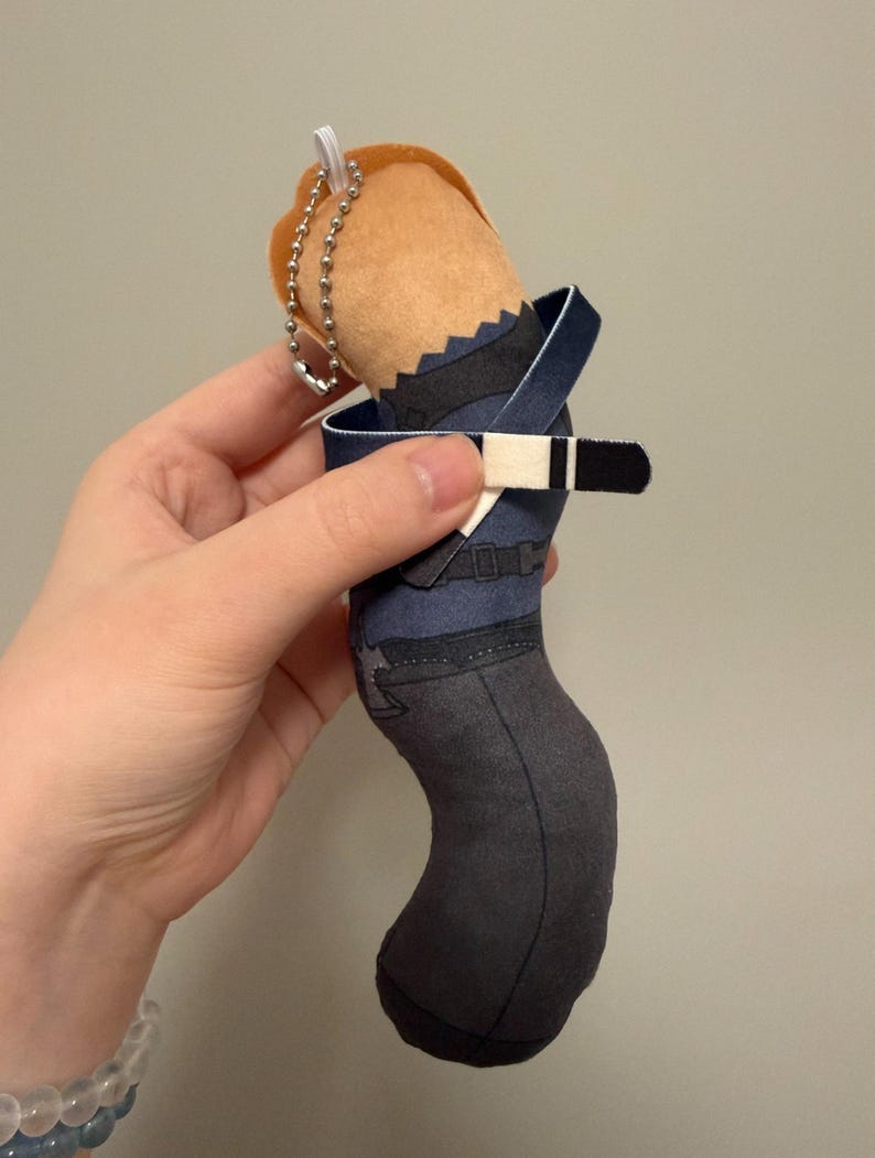 May include: A plush character keychain featuring a tan head with orange hair, a dark blue shirt with a black belt, and dark gray pants. The figure includes a silver metal ball chain attached to the top for hanging, with a printed fabric design.