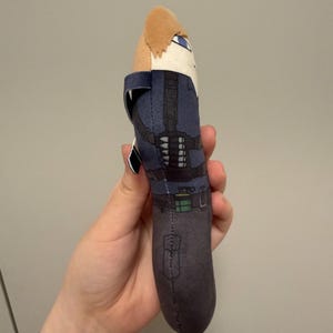 May include: A plush toy shaped like a character with light brown hair, wearing a blue tactical vest over a dark shirt, black pants, and a belt with a green pouch. The figure has a printed design showing a harness and gear on the torso.