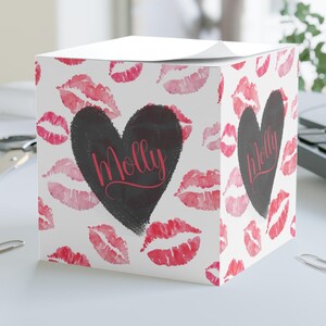 以下が含まれることがあります： 赤い口紅のキスの模様が入った白い立方体のメモ帳。前面中央には、ピンクの筆記体で「Molly」と書かれた黒いハートがあります。メモ帳は、ペーパークリップの付いた白い机の上にあります。