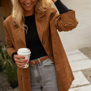 May include: A brown suede blazer, black top, and grey jeans. The blazer has two buttons and pockets. A brown braided belt with a gold buckle is worn with the jeans. A white coffee cup is held in the hand.