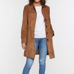 May include: A woman wearing a brown suede trench coat over a white top and blue jeans. The coat has a button-up closure and a classic collar. The jeans are light blue with a distressed detail.