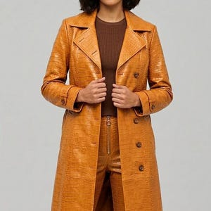 Könnte beinhalten: Eine Frau trägt einen langen, ockerfarbenen Trenchcoat aus Kunstleder mit Krokodilhautstruktur. Der Mantel hat ein doppelreihiges Design mit großen Knöpfen und einem Reverskragen. Die Frau trägt auch eine passende Hose und ein braunes Oberteil.