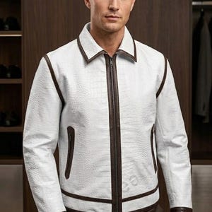 Könnte beinhalten: Weiße Lederjacke mit braunen Zierleisten und Reißverschluss. Die Jacke hat ein strukturiertes Muster und einen Kragen. Die Jacke wird mit braunen Hosen getragen. Die Jacke hat zwei Vordertaschen.
