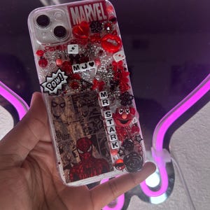 Può includere: Custodia trasparente per telefono decorata con abbellimenti a tema Marvel. La custodia presenta un fumetto di Spider-Man, il logo Marvel e vari ciondoli rossi e neri, tra cui labbra, cuori e dadi. È visibile anche la parola "POW!".