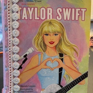 May include: A Little Golden Book biography of Taylor Swift. The cover features an illustration of Taylor Swift with blonde hair, forming a heart with her hands. The book is decorated with pink floral fabric and white lace trim.
