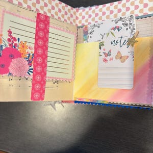 Pu&ograve; includere: Diario aperto con disegni floreali e farfalle. La pagina sinistra presenta una composizione floreale con fiori rosa e arancioni. La pagina destra ha una tasca con una carta che dice "note" e ha illustrazioni di farfalle. Un nastro washi rosa a fantasia &egrave; sulla pagina sinistra.