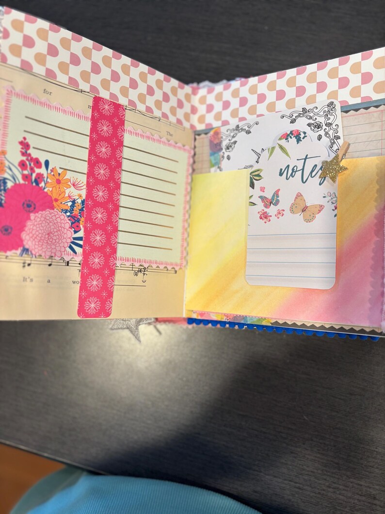 Pu&ograve; includere: Diario aperto con disegni floreali e farfalle. La pagina sinistra presenta una composizione floreale con fiori rosa e arancioni. La pagina destra ha una tasca con una carta che dice "note" e ha illustrazioni di farfalle. Un nastro washi rosa a fantasia &egrave; sulla pagina sinistra.