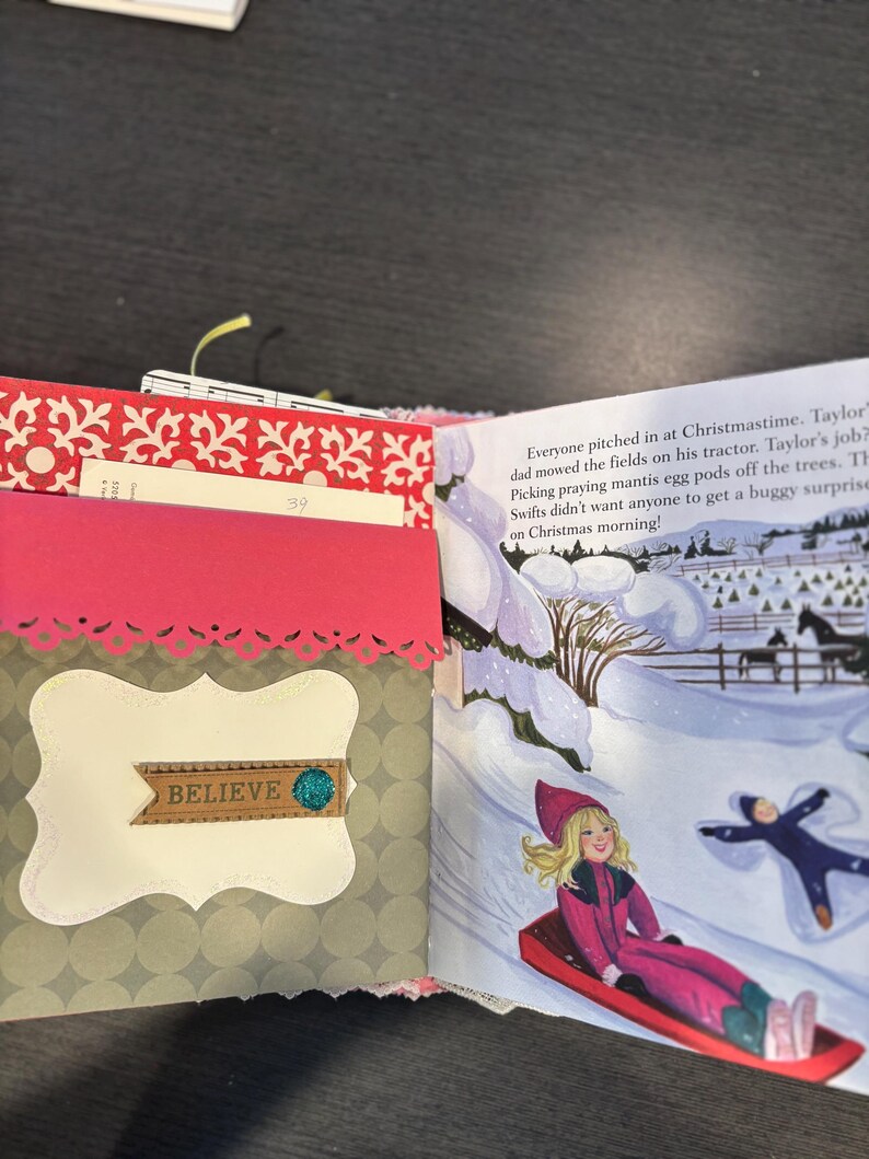 Pu&ograve; includere: Diario aperto con copertina fantasia rossa e bianca, con tasca rosa con bordo decorativo. La pagina sinistra ha un'etichetta decorativa con la parola "BELIEVE". La pagina destra mostra una scena invernale con bambini che vanno in slitta.