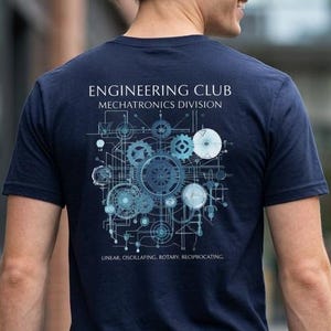 May include: A navy blue t-shirt featuring a white graphic on the back with the text ENGINEERING CLUB MECHATRONICS DIVISION and a complex mechanical gear design with the words LINEAR, OSCILLATING, ROTARY, RECIPROCATING printed below the gears.