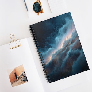 Könnte beinhalten: Spiralgebundenes Notizbuch mit einem Nebeldesign in Blau, Türkis und Gold. Das Notizbuch ist geöffnet und zeigt leere weiße Seiten. Eine goldene Büroklammer und eine Sonnenbrille sind ebenfalls sichtbar.