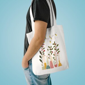 May include: White tote bag with a science-themed illustration. The design features colourful beakers, holly, and fairy lights. The bag is slung over a shoulder, displaying the festive artwork.