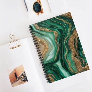 Könnte beinhalten: Spiralgebundenes Notizbuch mit einem grün-goldenen Marmordesign auf dem Einband. Das Notizbuch liegt offen auf einer weißen Oberfläche, mit einer goldenen Büroklammer und Sonnenbrille. Das Cover hat ein wirbelndes Muster mit Glitzerakzenten.