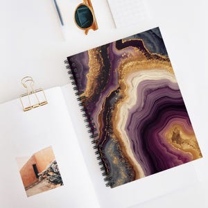 Könnte beinhalten: Spiralgebundenes Notizbuch mit einem Achat-Design in Lila, Gold und Creme. Das Notizbuch ist geöffnet und zeigt das farbenfrohe Muster auf der rechten Seite. Eine goldene Büroklammer und Sonnenbrille sind ebenfalls sichtbar.
