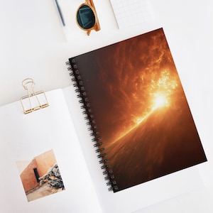 Könnte beinhalten: Spiralgebundenes Notizbuch mit einem lebendigen, feurigen abstrakten Design. Das Cover zeigt einen hellen orange-gelben Lichtausbruch vor dunklem Hintergrund. Das Notizbuch liegt offen auf einer weißen Oberfläche, mit einer goldenen Büroklammer und Sonnenbrille.