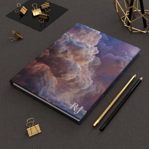 Op de afbeelding: Een hardcover notitieboek met een hemelse wolkenontwerp in paars, oranje en wit. De initialen "RJ" staan rechtsonder gedrukt. Gouden en zwarte potloden en gouden paperclips zijn ook zichtbaar.
