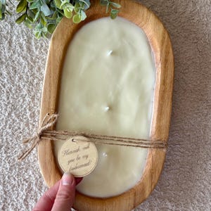 May include: A large, oval wooden candle holder filled with a creamy white candle. The candle has three wicks. A small wooden tag tied with twine reads "Hannah, will you be my bridesmaid?" Greenery is in the upper left corner.
