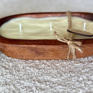 May include: A large, oval candle in a rustic wooden bowl. The candle is a creamy, off-white colour with three wicks. A wooden tag is tied to the bowl with twine. The bowl is brown with visible wood grain.