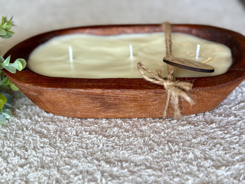 May include: A large, oval candle in a rustic wooden bowl. The candle is a creamy, off-white colour with three wicks. A wooden tag is tied to the bowl with twine. The bowl is brown with visible wood grain.
