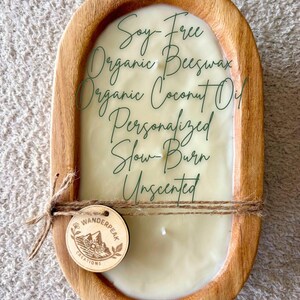 May include: A wooden candle holder with a cream-coloured candle. The candle is labelled "Soy-Free Organic Beeswax Organic Coconut Oil Personalized Slow-Burn Unscented." A small wooden tag with the Wanderpeak Creations logo is attached.