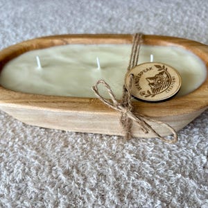 May include: An oval wooden dough bowl candle with three wicks. The candle wax is a light colour. A wooden tag with the text "Wanderpeak Creations" is tied with twine. The bowl sits on a textured surface.