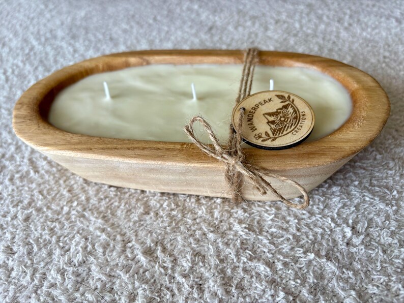 May include: An oval wooden dough bowl candle with three wicks. The candle wax is a light colour. A wooden tag with the text "Wanderpeak Creations" is tied with twine. The bowl sits on a textured surface.