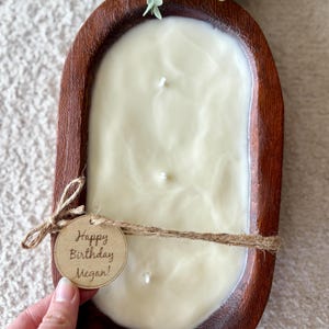 May include: A cream-coloured candle with three wicks, in a dark brown wooden bowl. The bowl is tied with twine and a tag that reads "Happy Birthday Megan!". Greenery is visible in the background.