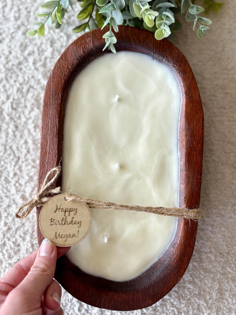 May include: A cream-coloured candle with three wicks, in a dark brown wooden bowl. The bowl is tied with twine and a tag that reads "Happy Birthday Megan!". Greenery is visible in the background.