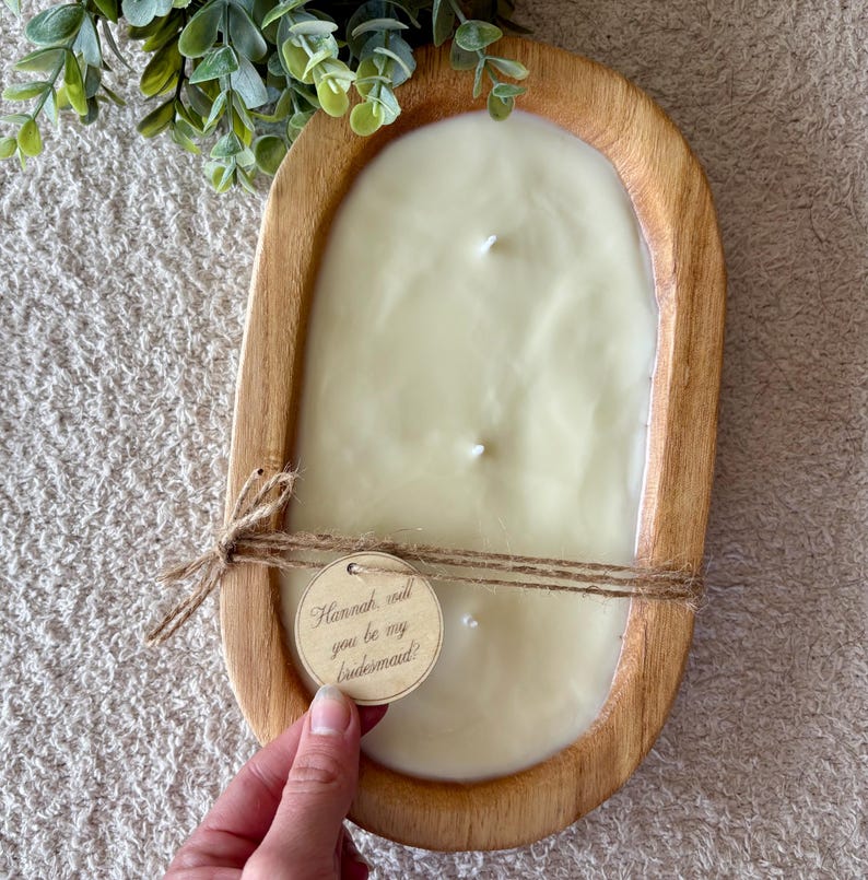 May include: A large, oval wooden candle holder filled with a creamy white candle. The candle has three wicks. A small wooden tag tied with twine reads "Hannah, will you be my bridesmaid?" Greenery is in the upper left corner.