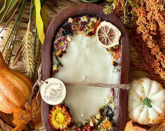 May include: A cream-colored candle in a wooden oval container, decorated with dried flowers and a slice of dried citrus. The candle is tied with twine and a wooden tag. The image includes fall foliage, gourds, and a small pumpkin.