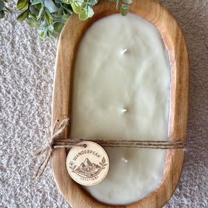 May include: An ivory-coloured candle with three wicks, set in an oval wooden bowl. The bowl has a natural wood finish. A wooden tag with the text "Wanderpeak Creations" is tied with twine. Green foliage is visible.