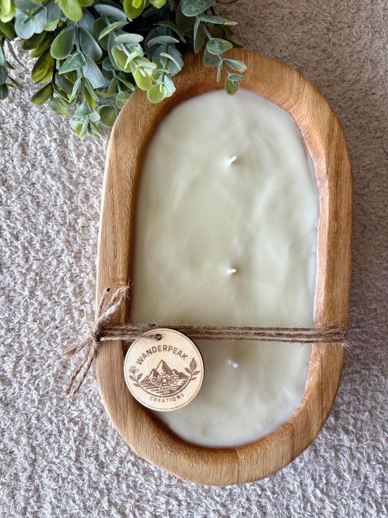 May include: An ivory-coloured candle with three wicks, set in an oval wooden bowl. The bowl has a natural wood finish. A wooden tag with the text "Wanderpeak Creations" is tied with twine. Green foliage is visible.
