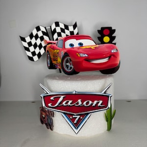 May include: A red Lightning McQueen car cake topper with the name "Jason" and the number "7". The topper includes a checkered flag, a traffic light, and a cactus. The car has the number 95 on it.