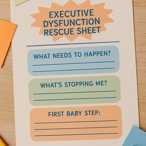 May include: A cream-colored paper titled "EXECUTIVE DYSFUNCTION RESCUE SHEET" with sections for "WHAT NEEDS TO HAPPEN?", "WHAT'S STOPPING ME?", and "FIRST BABY STEP:". The paper is on a wooden surface with sticky notes and a pencil.