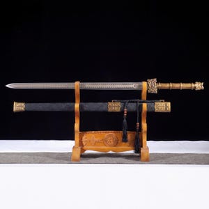 Puede incluir: Una espada de doble filo con hoja de acero estampada y empuñadura de metal dorado, apoyada sobre un soporte de madera encima de su vaina negra. La vaina presenta herrajes de metal dorado y una borla negra con cuentas de madera marrón.