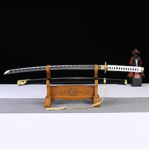 May include: A katana sword with a silver blade featuring a wavy hamon line, a white wrapped handle with diamond-shaped cutouts, and a gold-colored guard. The black scabbard rests on a wooden stand with carved details and a hanging beige cord.