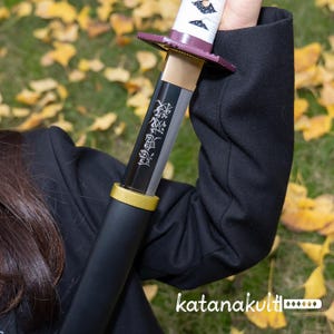 May include: A black katana with a gold hilt and a purple guard. The blade has Japanese characters etched on it. The handle is white with a geometric design. The text "katanakult" is visible at the bottom.