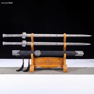 May include: Two silver swords with ornate handles and blades are displayed on a wooden stand. A black scabbard with silver accents rests below. The stand has a carved design and tassels.