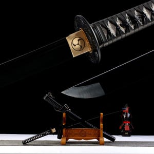 Pode incluir: Espada katana preta com um emblema dourado na guarda. A empunhadura é envolta em material preto. A lâmina é preta e reflete a luz. A espada é exibida em um suporte de madeira com uma figura de samurai em miniatura.