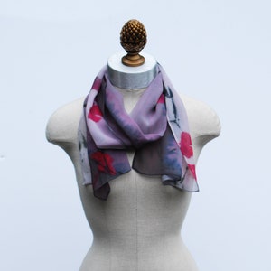 May include: A gray silk scarf with a floral print in shades of pink and red. The scarf is draped around a mannequin's neck.