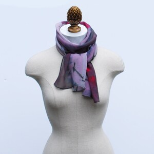May include: A purple and red silk scarf with a painterly design. The scarf is tied around a mannequin's neck.