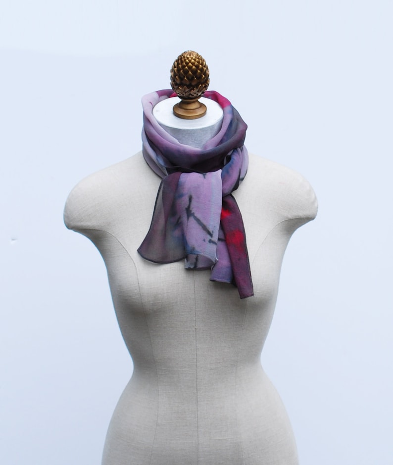 May include: A purple and red silk scarf with a painterly design. The scarf is tied around a mannequin's neck.