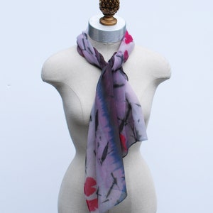 May include: A purple and pink silk scarf with black and red floral designs. The scarf is tied around a mannequin's neck.