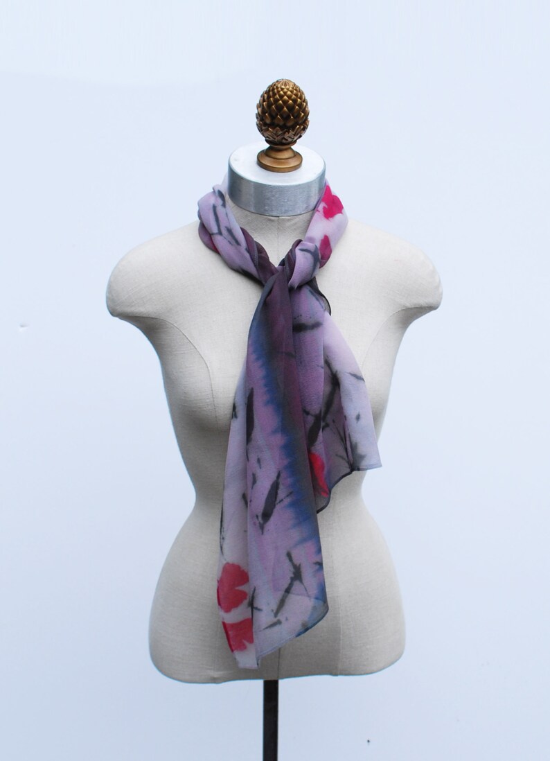 May include: A purple and pink silk scarf with black and red floral designs. The scarf is tied around a mannequin's neck.