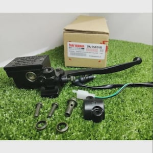 Op de afbeelding: Een zwarte motorfiets remhoofdcilinder met hendel, vloeistofreservoir en kabelboom. De set bevat twee metalen bouten, twee ringen en een kleine zwarte montagebeugel, alles verpakt met een Thai Yamaha 2RJ-25870-00 doos.