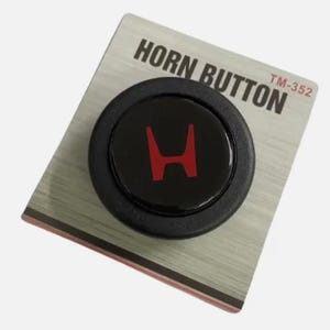以下が含まれることがあります： 赤い「H」ロゴが付いた黒いホーンボタン。銀色のカードに装着されています。カードには「HORN BUTTON」と「TM-352」の文字が印刷されています。ボタンは丸く、光沢のある仕上がりです。