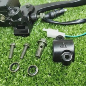 Op de afbeelding: Een zwarte motorfiets remcilinder-assemblage met een hendel, een witte elektrische connector met een groene draad, twee kleine metalen bouten, een banjobout, twee ronde ringen en een zwarte montagebeugel met het woord UP in reliëf.