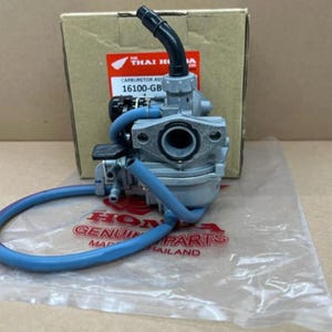 May include: A silver metal carburettor assembly featuring a black plastic elbow fitting and a blue rubber hose. The unit is positioned in front of a cardboard box with a label reading FOR THAI HONDA and 16100-GB, resting on a clear plastic bag.