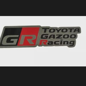 May include: A rectangular emblem with the text "TOYOTA GAZOO Racing". The emblem has a grey border with black and red accents. The letters "GR" are displayed in red within a black and grey section.