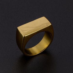 Puede incluir: Un anillo de sello dorado con una parte superior rectangular. El anillo tiene una superficie lisa y pulida y una banda gruesa. El anillo se muestra sobre un fondo oscuro.