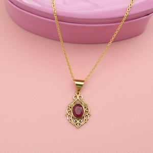 Puede incluir: Un collar dorado con un colgante con una piedra preciosa roja ovalada. El colgante está engastado en un marco ornamentado de estilo filigrana. El collar se exhibe sobre una superficie rosa.
